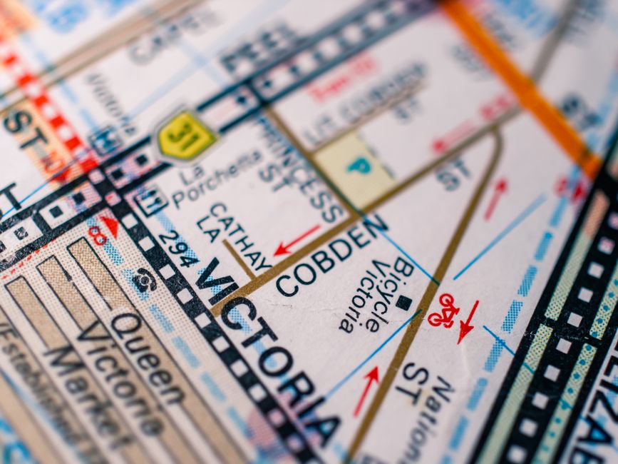 A close-up photograph of a detailed map showing the streets and nearby area around Cobden in Queens Park. The map highlights surrounding roads, with street names such as Queen, Vitoria, and Cathay, along with symbols indicating bicycle lanes and traffic directions. The map features a variety of colors, including white for the background, black for street boundaries, blue for water features or transportation routes, red for traffic arrows, and yellow for directional or signage markers. The texture appears smooth with clear printed lines and labels, and the focus emphasizes the central section around Cobden, with other streets and details fading into the background. This detailed map could be used to understand local navigation or planning for independent or private waste removal services, such as those provided by House Clearance Queens Park, especially in the context of managing rubbish collection or property clearance in the area, supporting environmentally conscious waste handling.