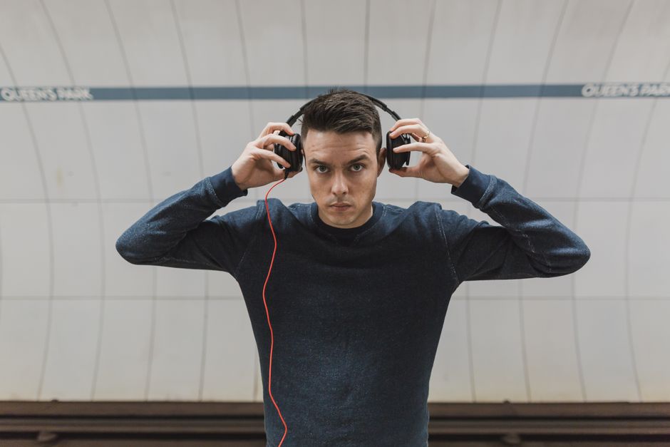 A young man with short dark hair and a serious expression is standing indoors against a curved, light-colored tiled wall with blue lines and text. He is wearing a dark long-sleeved shirt and is adjusting large black over-ear headphones with both hands, with a red audio cable hanging down. The environment suggests a modern indoor space such as a subway station or public transport hub, with the background featuring a tiled surface and lighting that creates minimal shadows. This scene is visually related to personal audio equipment, which could be associated with temporary arrangements or private listening experiences, indirectly connected to the context of waste management or rubbish removal services like those offered by House Clearance Queens Park, though the image itself does not depict rubbish or removal activities.