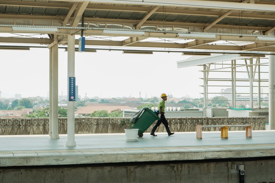 A worker wearing a yellow safety helmet and a green high-visibility vest is walking along an outdoor station platform, pulling a large green wheelie bin with black wheels. The bin is positioned slightly behind a white plastic bucket placed on the paved surface of the platform. The platform is sheltered by a metal roof supported by white columns and features a metallic railing with decorative stonework underneath it, separating the platform from the view beyond. In the background, a cityscape with low-rise buildings, rooftops, and trees is visible under overcast or diffused lighting, creating a neutral atmosphere that suggests ongoing maintenance or waste collection activity. The scene reflects the environment of a public transport station involved in or adjacent to waste collection or disposal operations, aligned with private or independent rubbish removal services as commonly provided by companies like House Clearance Queens Park.