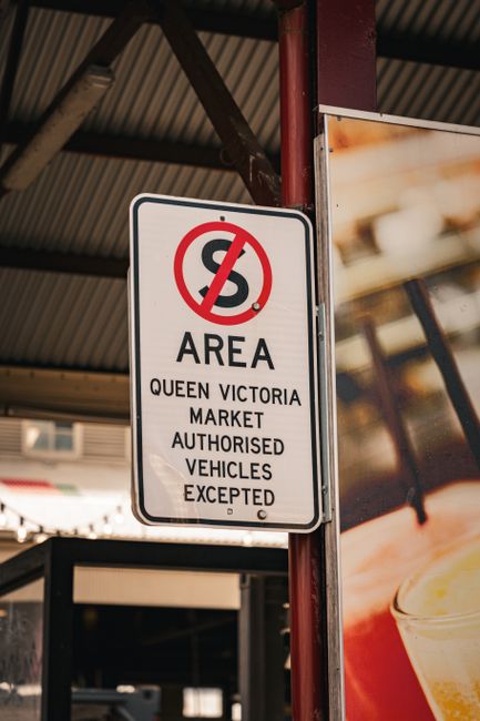 A rectangular white sign with a black border, mounted on a red metal post under a large, grey corrugated roof structure. The sign features a red circle with a diagonal line crossing out the letter 'S,' indicating restrictions, above the text that reads 'AREA QUEEN VICTORIA MARKET AUTHORISED VEHICLES EXCEPTED.' The background includes part of a building with windows, and a blurred partial view of a brightly colored poster or advertisement, suggesting a busy commercial or market environment. The lighting appears natural, with no significant shadows or reflections. This scene relates to designated zones where specific vehicle access is regulated, fitting within the context of managing waste removal or delivery access at commercial sites, as seen in some private or independent collection arrangements that do not follow local authority vehicle permissions.